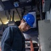 A young male technician or worker wearing a blue hard hat and safety glasses is examining a document or checklist while standing next to a metal electrical or equipment panel. He is dressed in dark navy blue coveralls and appears to be in an industrial or mechanical workspace. An orange ladder is visible in the background among various cables, equipment, and overhead infrastructure.