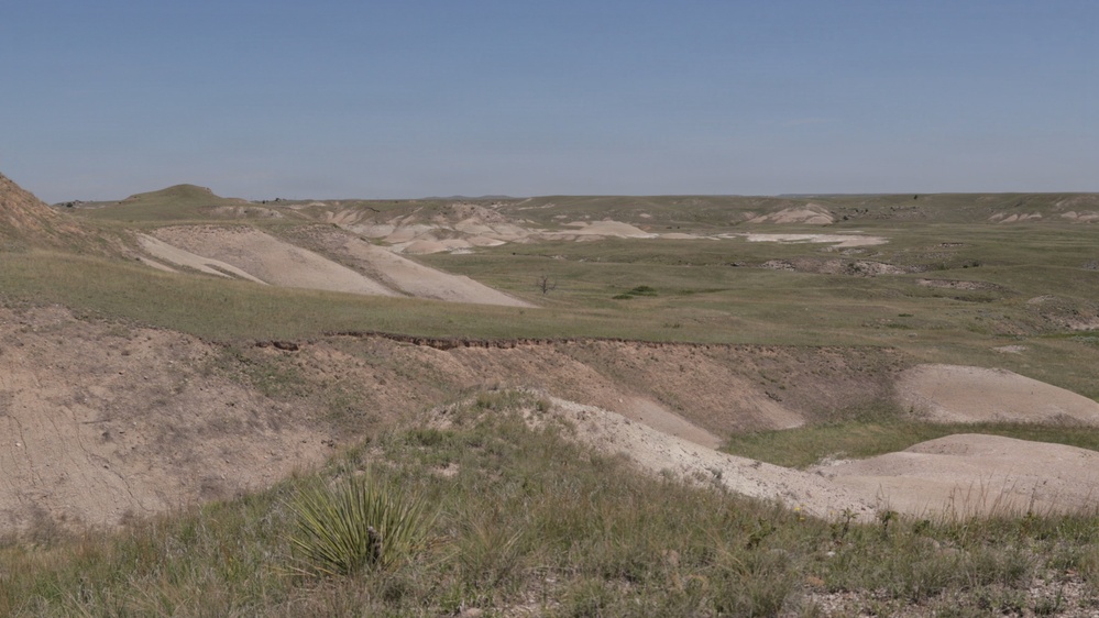 A wide-angle landscape photograph depicting an arid to semi-arid badlands terrain with rolling eroded hills and sparse grassland vegetation. The scene shows a mix of exposed sandy/clay soils and patchy green grass cover under a clear blue sky. The terrain is characteristic of the Great Plains badlands region, possibly South Dakota or Nebraska.