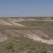 A wide-angle landscape photograph depicting an arid to semi-arid badlands terrain with rolling eroded hills and sparse grassland vegetation. The scene shows a mix of exposed sandy/clay soils and patchy green grass cover under a clear blue sky. The terrain is characteristic of the Great Plains badlands region, possibly South Dakota or Nebraska.