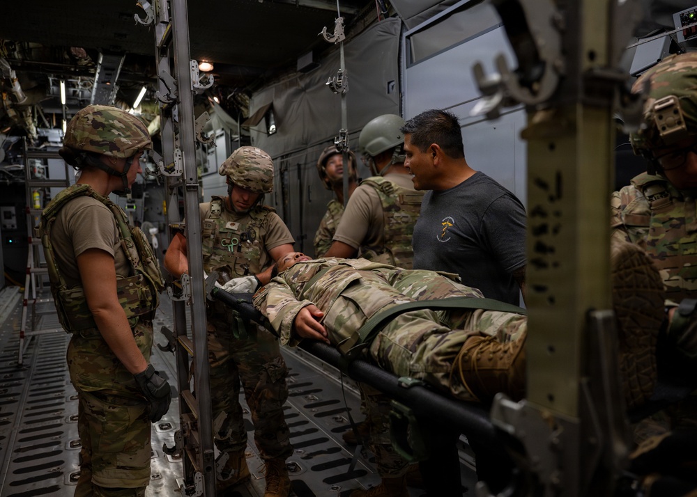 Military personnel are conducting what appears to be a medical evacuation (MEDEVAC) training exercise or actual casualty transport inside a military aircraft, likely a C-130 or similar transport aircraft. Several soldiers in multicam/OCP uniforms and combat helmets are handling a person on a litter/stretcher. One individual in civilian clothing (gray t-shirt) is also present and appears to be observing or assisting.