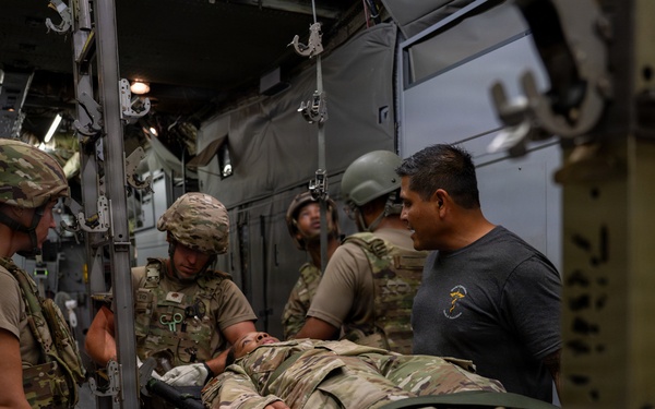 Military personnel are conducting what appears to be a medical evacuation (MEDEVAC) training exercise or actual casualty transport inside a military aircraft, likely a C-130 or similar transport aircraft. Several soldiers in multicam/OCP uniforms and combat helmets are handling a person on a litter/stretcher. One individual in civilian clothing (gray t-shirt) is also present and appears to be observing or assisting.