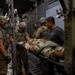 Military personnel are conducting what appears to be a medical evacuation (MEDEVAC) training exercise or actual casualty transport inside a military aircraft, likely a C-130 or similar transport aircraft. Several soldiers in multicam/OCP uniforms and combat helmets are handling a person on a litter/stretcher. One individual in civilian clothing (gray t-shirt) is also present and appears to be observing or assisting.