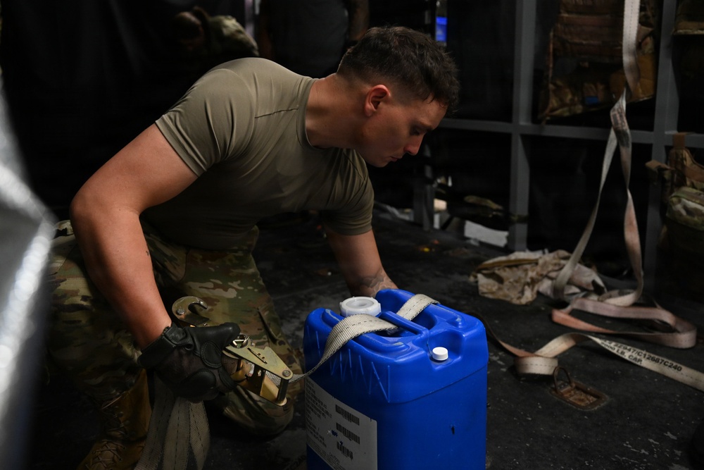 A military service member in OCP (Operational Camouflage Pattern) uniform and black tactical gloves is securing a blue plastic jerry can or water container using a ratchet strap. The scene appears to be inside a military aircraft cargo hold or transport vehicle, with cargo straps and equipment visible in the background. The soldier is focused on tightening or fastening the restraint strap around the container.