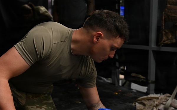 A military service member in OCP (Operational Camouflage Pattern) uniform and black tactical gloves is securing a blue plastic jerry can or water container using a ratchet strap. The scene appears to be inside a military aircraft cargo hold or transport vehicle, with cargo straps and equipment visible in the background. The soldier is focused on tightening or fastening the restraint strap around the container.