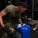 A military service member in OCP (Operational Camouflage Pattern) uniform and black tactical gloves is securing a blue plastic jerry can or water container using a ratchet strap. The scene appears to be inside a military aircraft cargo hold or transport vehicle, with cargo straps and equipment visible in the background. The soldier is focused on tightening or fastening the restraint strap around the container.