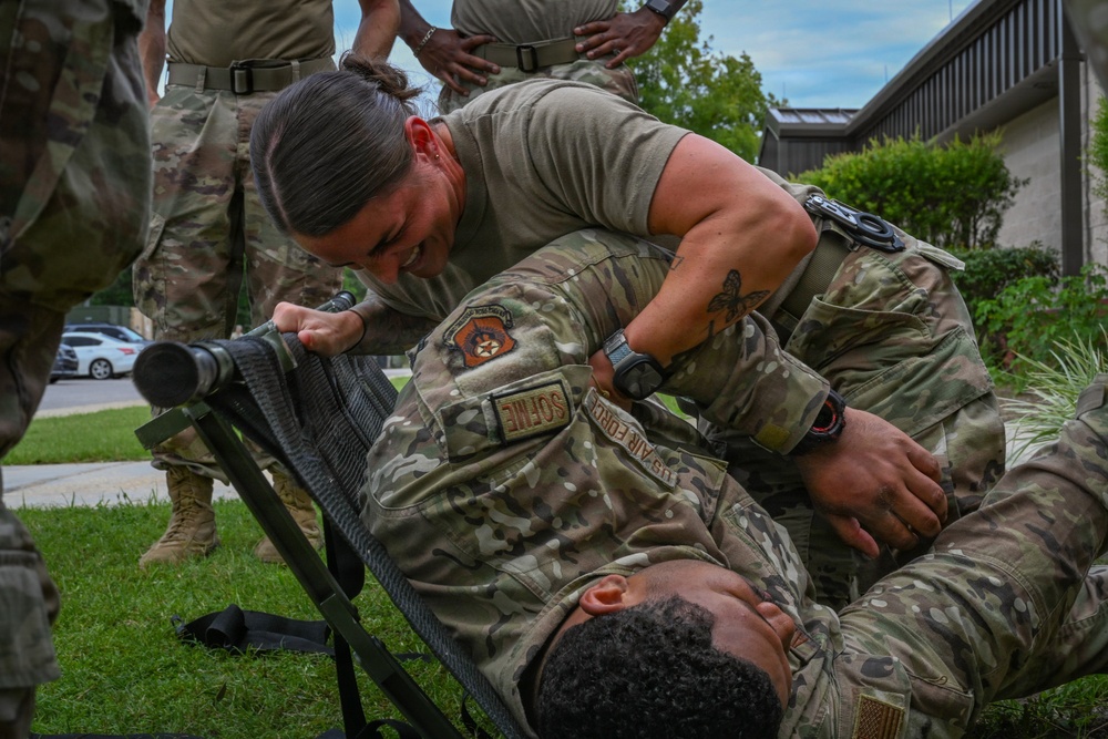 A US Air Force training exercise is underway outdoors on a grassy area, where one service member is practicing casualty care or litter-carry techniques on another who is lying on a military stretcher. The standing individual wears OCP (Operational Camouflage Pattern) uniform with visible 'US AIR FORCE' and 'SOFME' (Special Operations Forces Medical Element) tape and a unit patch. Several other uniformed personnel observe in the background.