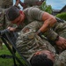 A US Air Force training exercise is underway outdoors on a grassy area, where one service member is practicing casualty care or litter-carry techniques on another who is lying on a military stretcher. The standing individual wears OCP (Operational Camouflage Pattern) uniform with visible 'US AIR FORCE' and 'SOFME' (Special Operations Forces Medical Element) tape and a unit patch. Several other uniformed personnel observe in the background.