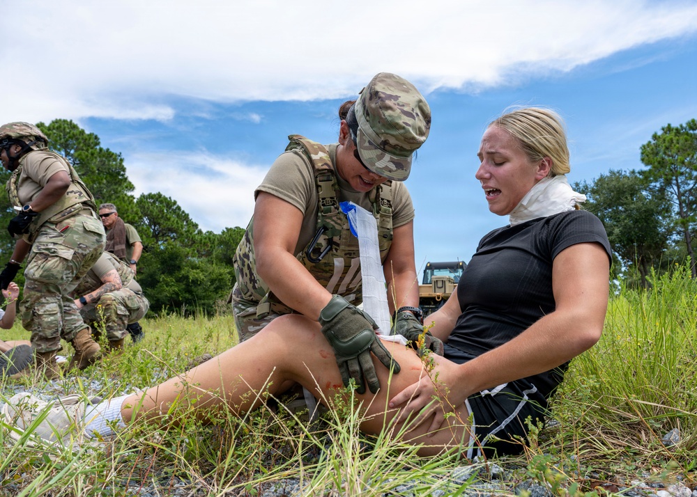 A military medic in full combat uniform and tactical vest is providing first aid to a civilian woman seated on the ground, applying a bandage or dressing to her leg. The scene appears to be a training exercise or field medical drill conducted in an outdoor grassy area. Additional military personnel are visible in the background engaged in similar activities.