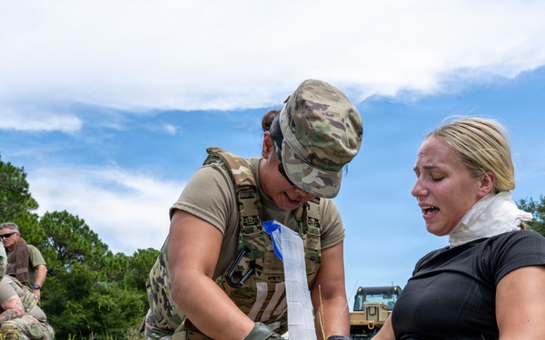 A military medic in full combat uniform and tactical vest is providing first aid to a civilian woman seated on the ground, applying a bandage or dressing to her leg. The scene appears to be a training exercise or field medical drill conducted in an outdoor grassy area. Additional military personnel are visible in the background engaged in similar activities.