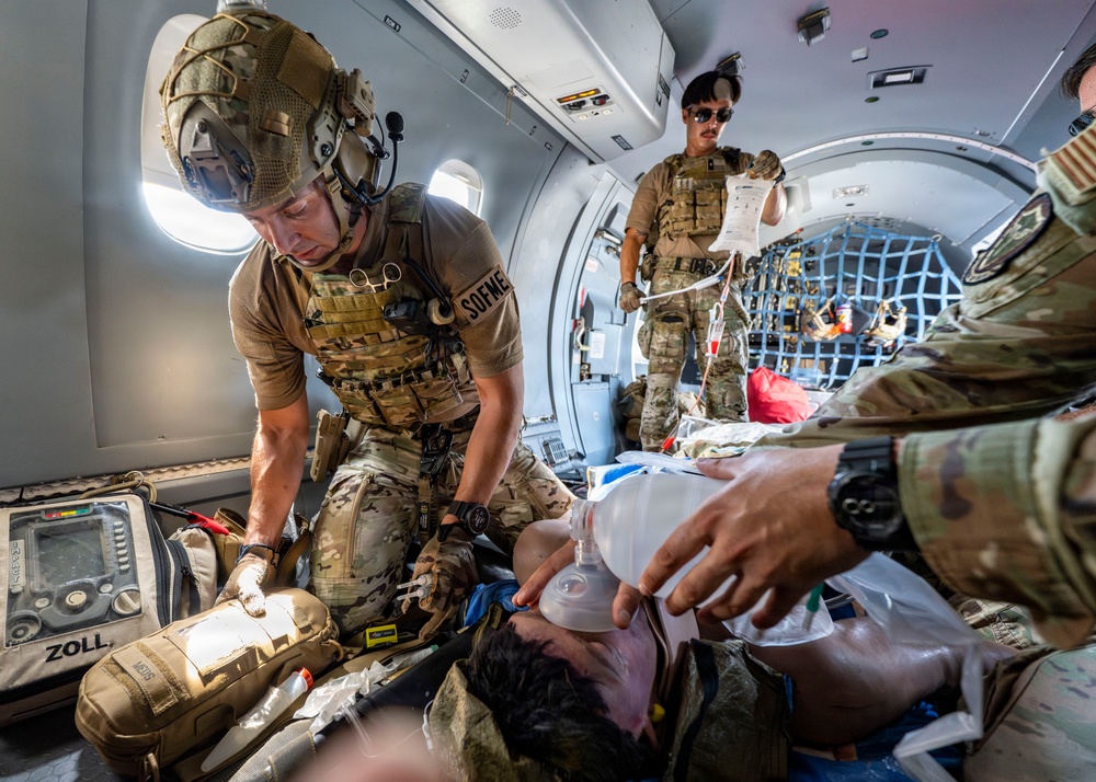 Military medical personnel (SOF medics) are providing emergency care to a casualty inside what appears to be a military aircraft. At least three uniformed personnel in MultiCam/OCP camouflage are actively treating the patient, with one administering a bag-valve mask for airway management and another monitoring IV fluids. A ZOLL medical device (defibrillator/monitor) is visible alongside other medical equipment.