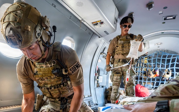 Military medical personnel (SOF medics) are providing emergency care to a casualty inside what appears to be a military aircraft. At least three uniformed personnel in MultiCam/OCP camouflage are actively treating the patient, with one administering a bag-valve mask for airway management and another monitoring IV fluids. A ZOLL medical device (defibrillator/monitor) is visible alongside other medical equipment.