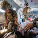Military medical personnel (SOF medics) are providing emergency care to a casualty inside what appears to be a military aircraft. At least three uniformed personnel in MultiCam/OCP camouflage are actively treating the patient, with one administering a bag-valve mask for airway management and another monitoring IV fluids. A ZOLL medical device (defibrillator/monitor) is visible alongside other medical equipment.
