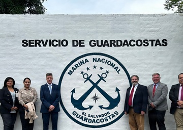 A group of seven individuals poses in front of a white wall bearing the insignia and text of the 'Servicio de Guardacostas' (Coast Guard Service) of El Salvador's Marina Nacional. The group appears to include both military and civilian personnel, suggesting an official visit or diplomatic meeting. The setting is an outdoor facility associated with the El Salvador National Navy Coast Guard.