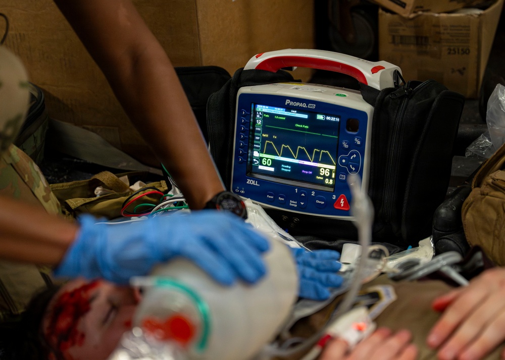 A medical emergency scenario showing a ZOLL Propaq MD portable patient monitor displaying vital signs, with gloved hands performing patient care. The monitor shows a heart rate of 60 BPM and SpO2 of 96%, with a yellow waveform visible on screen. Personnel wearing blue nitrile gloves and military-pattern clothing are attending to a patient with visible simulated or real blood present.