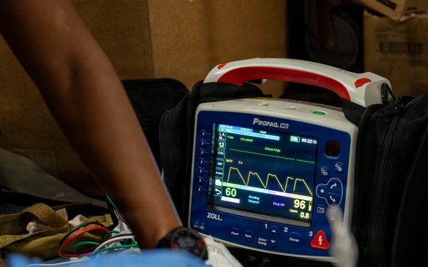 A medical emergency scenario showing a ZOLL Propaq MD portable patient monitor displaying vital signs, with gloved hands performing patient care. The monitor shows a heart rate of 60 BPM and SpO2 of 96%, with a yellow waveform visible on screen. Personnel wearing blue nitrile gloves and military-pattern clothing are attending to a patient with visible simulated or real blood present.