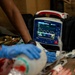 A medical emergency scenario showing a ZOLL Propaq MD portable patient monitor displaying vital signs, with gloved hands performing patient care. The monitor shows a heart rate of 60 BPM and SpO2 of 96%, with a yellow waveform visible on screen. Personnel wearing blue nitrile gloves and military-pattern clothing are attending to a patient with visible simulated or real blood present.