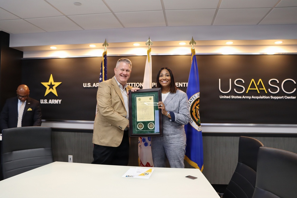 Two individuals are posing together in a formal setting at the United States Army Acquisition Support Center (USAASC), holding a framed certificate or award with two medallions. The ceremony appears to be a recognition or award presentation, taking place in an official conference room with U.S. Army branding. A third individual is visible in the background to the left.