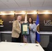 Two individuals are posing together in a formal setting at the United States Army Acquisition Support Center (USAASC), holding a framed certificate or award with two medallions. The ceremony appears to be a recognition or award presentation, taking place in an official conference room with U.S. Army branding. A third individual is visible in the background to the left.