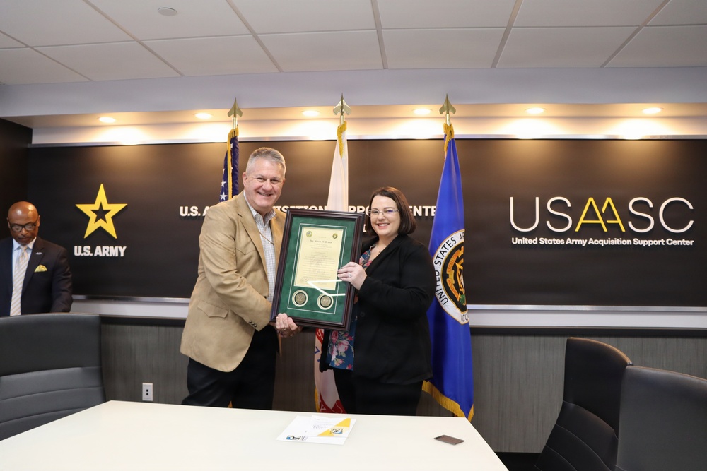 Two individuals are posing together while jointly holding a framed certificate or award at the United States Army Acquisition Support Center (USAASC). The setting appears to be an official ceremony or recognition event, with U.S. Army branding and flags prominently displayed in the background. A third individual is partially visible in the background to the left.