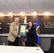 Two individuals are posing together while jointly holding a framed certificate or award at the United States Army Acquisition Support Center (USAASC). The setting appears to be an official ceremony or recognition event, with U.S. Army branding and flags prominently displayed in the background. A third individual is partially visible in the background to the left.