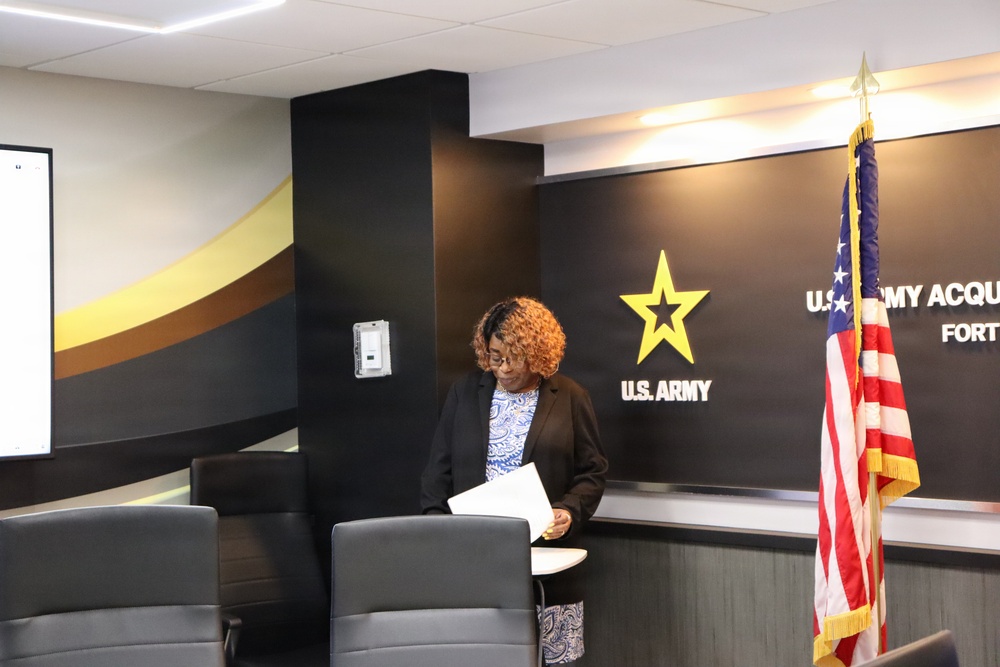 A woman stands at a podium or lectern reviewing papers in what appears to be a U.S. Army Acquisition Command conference or briefing room. She is dressed in a black blazer over a patterned blouse and has curly auburn hair. The room features the U.S. Army star logo, an American flag with gold fringe, and modern interior design with black and gold accents.