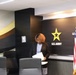 A woman stands at a podium or lectern reviewing papers in what appears to be a U.S. Army Acquisition Command conference or briefing room. She is dressed in a black blazer over a patterned blouse and has curly auburn hair. The room features the U.S. Army star logo, an American flag with gold fringe, and modern interior design with black and gold accents.