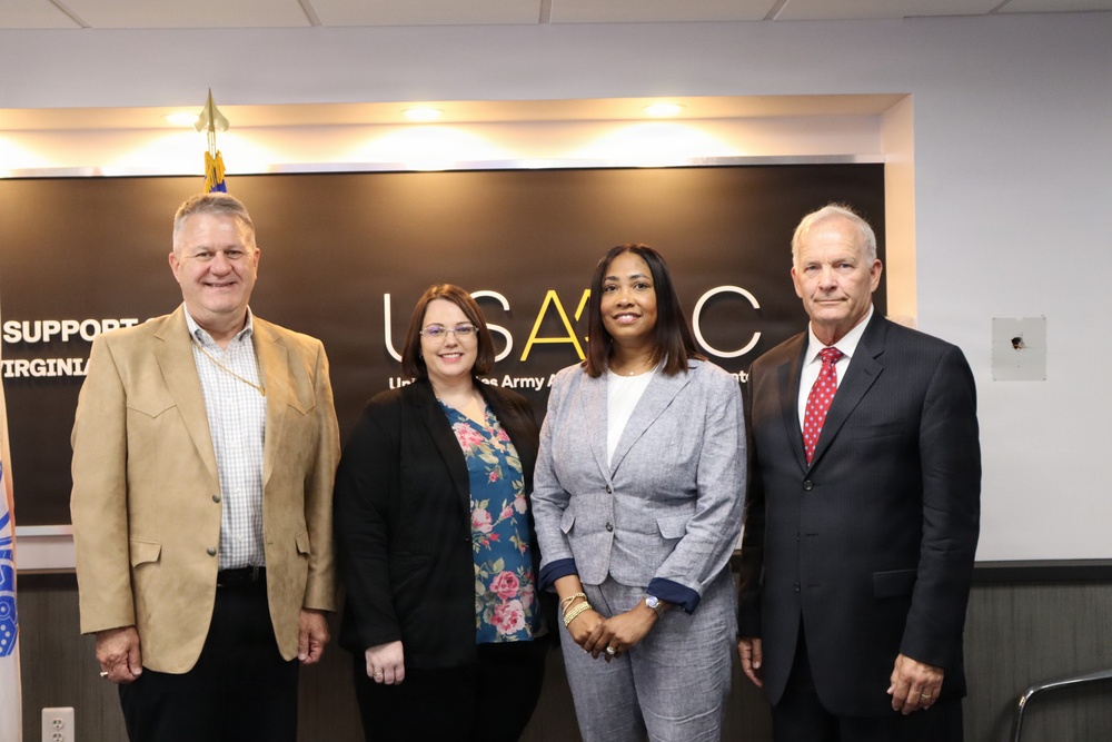 Four individuals are posing for a formal group photo in front of a USAASC (United States Army Acquisition Support Center) branded backdrop. The setting appears to be an official government or military-affiliated office environment. The group consists of two men and two women dressed in professional business attire.