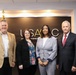 Four individuals are posing for a formal group photo in front of a USAASC (United States Army Acquisition Support Center) branded backdrop. The setting appears to be an official government or military-affiliated office environment. The group consists of two men and two women dressed in professional business attire.