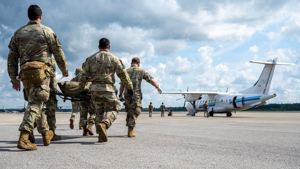 Military personnel in camouflage uniforms are carrying a patient on a litter/stretcher across an airfield tarmac toward a white twin-turboprop aircraft. The scene depicts a medical evacuation (MEDEVAC) operation, with multiple soldiers moving urgently toward the waiting aircraft. Additional personnel are visible near the aircraft in the background.