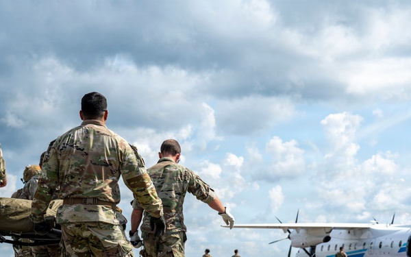 Military personnel in camouflage uniforms are carrying a patient on a litter/stretcher across an airfield tarmac toward a white twin-turboprop aircraft. The scene depicts a medical evacuation (MEDEVAC) operation, with multiple soldiers moving urgently toward the waiting aircraft. Additional personnel are visible near the aircraft in the background.