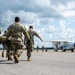 Military personnel in camouflage uniforms are carrying a patient on a litter/stretcher across an airfield tarmac toward a white twin-turboprop aircraft. The scene depicts a medical evacuation (MEDEVAC) operation, with multiple soldiers moving urgently toward the waiting aircraft. Additional personnel are visible near the aircraft in the background.