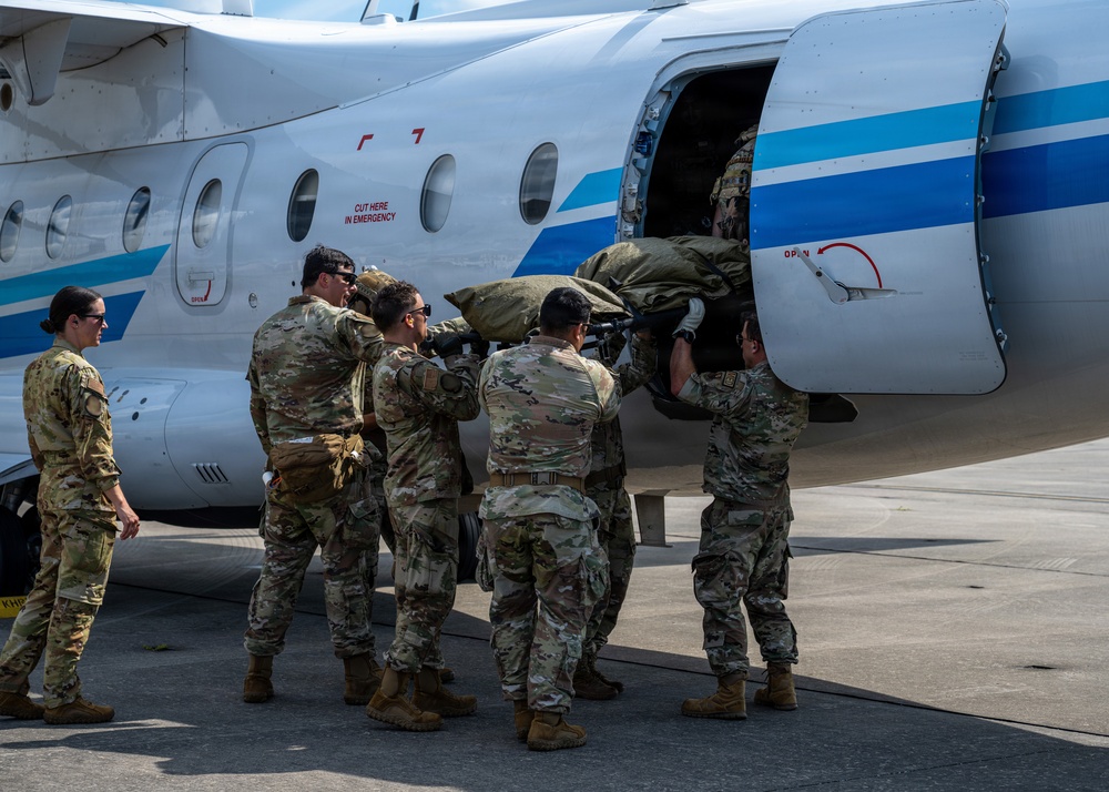 A group of military personnel in MultiCam/OCP camouflage uniforms are loading a large olive-drab bag or litter into the side cargo/passenger door of a small white civilian-style aircraft with blue stripe markings. One service member observes from the left while several others physically lift and push the load through the aircraft door. The scene takes place on a paved tarmac in what appears to be a warm-weather location.