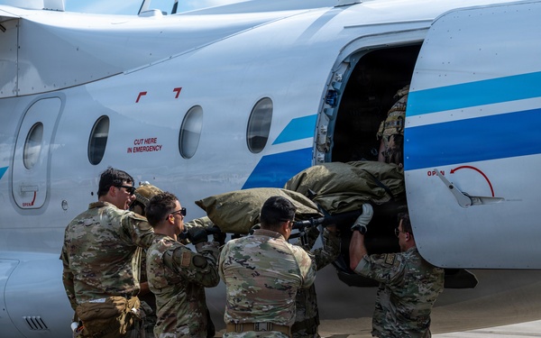 A group of military personnel in MultiCam/OCP camouflage uniforms are loading a large olive-drab bag or litter into the side cargo/passenger door of a small white civilian-style aircraft with blue stripe markings. One service member observes from the left while several others physically lift and push the load through the aircraft door. The scene takes place on a paved tarmac in what appears to be a warm-weather location.