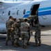 A group of military personnel in MultiCam/OCP camouflage uniforms are loading a large olive-drab bag or litter into the side cargo/passenger door of a small white civilian-style aircraft with blue stripe markings. One service member observes from the left while several others physically lift and push the load through the aircraft door. The scene takes place on a paved tarmac in what appears to be a warm-weather location.