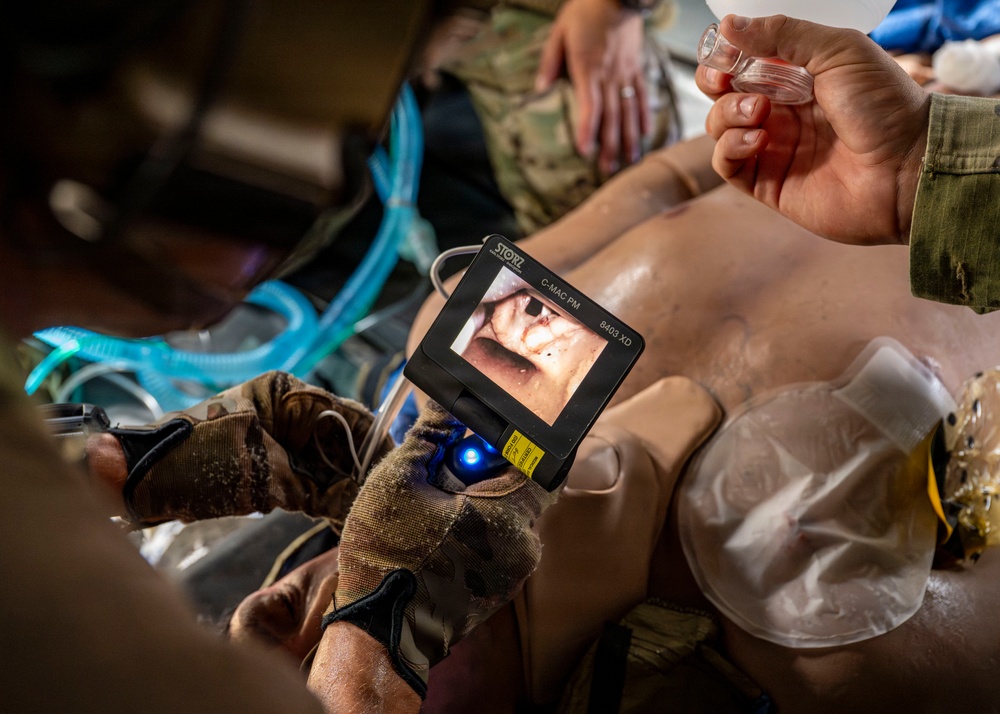 A military medical training exercise is being conducted on a medical mannequin. A person wearing tactical gloves is using a STORZ C-MAC PM video laryngoscope (model 8403 XD) to practice intubation on the training dummy. A second person is holding what appears to be an endotracheal tube or airway device, while a blue breathing circuit is visible nearby.