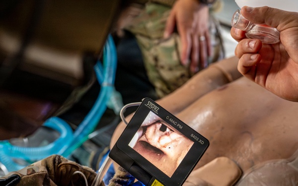 A military medical training exercise is being conducted on a medical mannequin. A person wearing tactical gloves is using a STORZ C-MAC PM video laryngoscope (model 8403 XD) to practice intubation on the training dummy. A second person is holding what appears to be an endotracheal tube or airway device, while a blue breathing circuit is visible nearby.