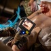 A military medical training exercise is being conducted on a medical mannequin. A person wearing tactical gloves is using a STORZ C-MAC PM video laryngoscope (model 8403 XD) to practice intubation on the training dummy. A second person is holding what appears to be an endotracheal tube or airway device, while a blue breathing circuit is visible nearby.