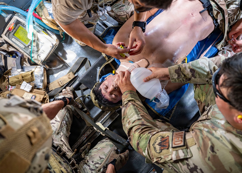 Military medical personnel are providing emergency care to a casualty lying on a litter/stretcher. Multiple providers in OCP (Operational Camouflage Pattern) uniforms are simultaneously performing airway management with a bag-valve mask and administering medications or IV access. Medical equipment including a ZOLL monitor/defibrillator and tactical medical gear are visible in the confined space, suggesting an aircraft or vehicle environment.