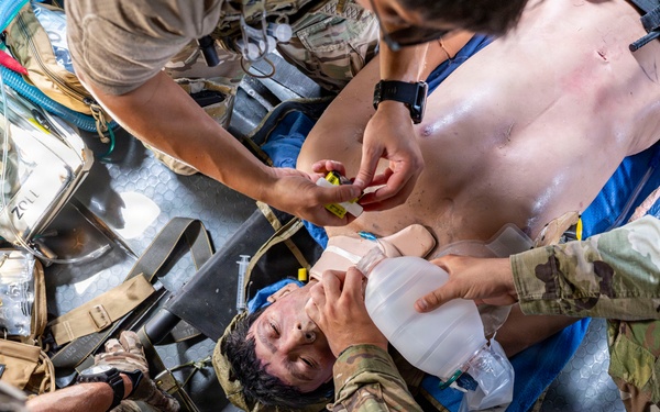 Military medical personnel are providing emergency care to a casualty lying on a litter/stretcher. Multiple providers in OCP (Operational Camouflage Pattern) uniforms are simultaneously performing airway management with a bag-valve mask and administering medications or IV access. Medical equipment including a ZOLL monitor/defibrillator and tactical medical gear are visible in the confined space, suggesting an aircraft or vehicle environment.
