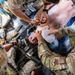 Military medical personnel are providing emergency care to a casualty lying on a litter/stretcher. Multiple providers in OCP (Operational Camouflage Pattern) uniforms are simultaneously performing airway management with a bag-valve mask and administering medications or IV access. Medical equipment including a ZOLL monitor/defibrillator and tactical medical gear are visible in the confined space, suggesting an aircraft or vehicle environment.