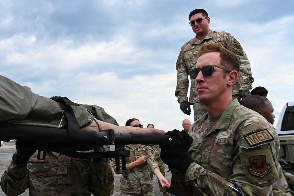 U.S. Air Force medical personnel are conducting what appears to be a casualty evacuation or aeromedical training exercise on a flight line or tarmac. Multiple airmen in OCP (Operational Camouflage Pattern) uniforms are handling a litter (stretcher) with a simulated or actual patient strapped to it. The prominent airman in the foreground wears a 'MED' patch, identifying him as medical personnel.