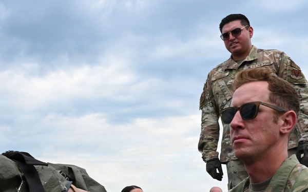 U.S. Air Force medical personnel are conducting what appears to be a casualty evacuation or aeromedical training exercise on a flight line or tarmac. Multiple airmen in OCP (Operational Camouflage Pattern) uniforms are handling a litter (stretcher) with a simulated or actual patient strapped to it. The prominent airman in the foreground wears a 'MED' patch, identifying him as medical personnel.
