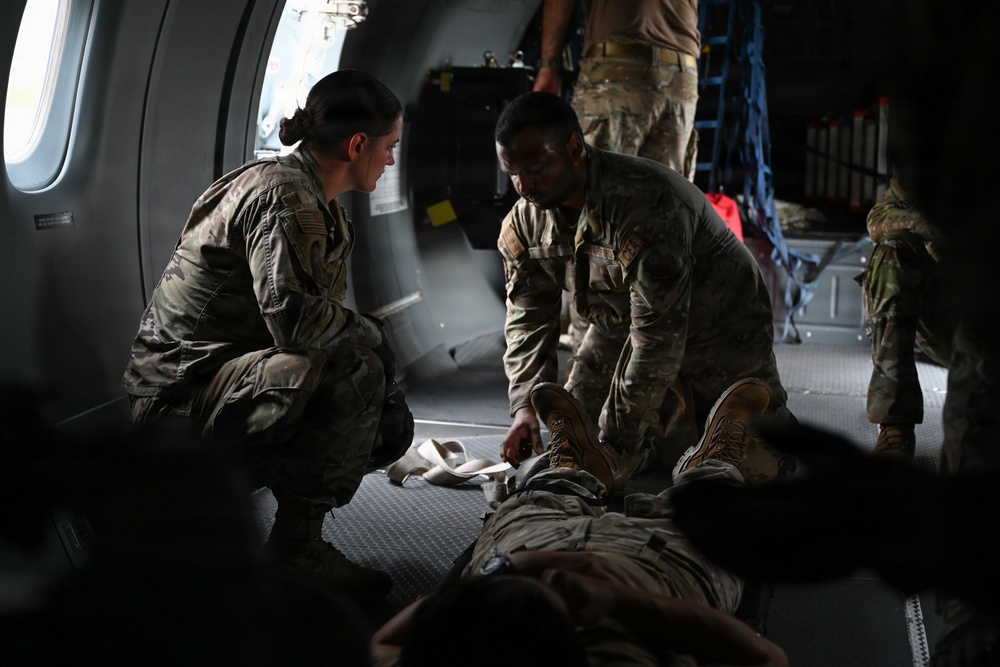 Military personnel in OCP (Operational Camouflage Pattern) uniforms are attending to a person lying on the floor inside what appears to be a military transport aircraft. Two soldiers are crouched over the individual on the ground, with one appearing to handle the person's legs or feet, suggesting a medical training exercise or casualty care scenario. Additional personnel are visible in the background of the aircraft interior.