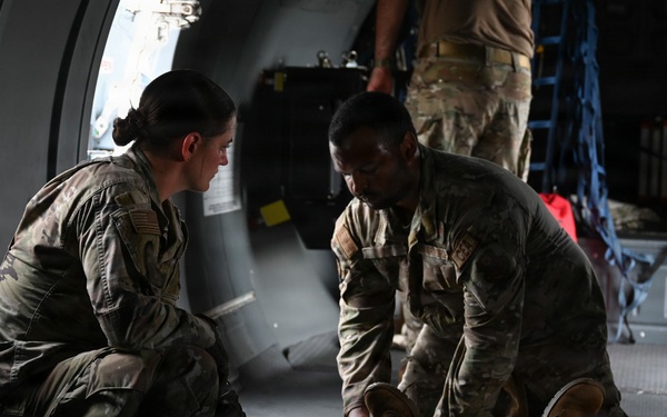 Military personnel in OCP (Operational Camouflage Pattern) uniforms are attending to a person lying on the floor inside what appears to be a military transport aircraft. Two soldiers are crouched over the individual on the ground, with one appearing to handle the person's legs or feet, suggesting a medical training exercise or casualty care scenario. Additional personnel are visible in the background of the aircraft interior.
