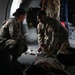 Military personnel in OCP (Operational Camouflage Pattern) uniforms are attending to a person lying on the floor inside what appears to be a military transport aircraft. Two soldiers are crouched over the individual on the ground, with one appearing to handle the person's legs or feet, suggesting a medical training exercise or casualty care scenario. Additional personnel are visible in the background of the aircraft interior.