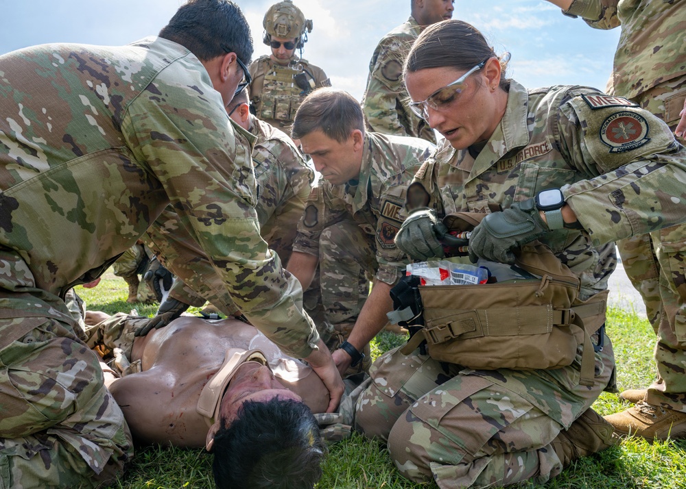 Multiple U.S. Air Force personnel in OCP (Operational Camouflage Pattern) uniforms are performing tactical combat casualty care (TCCC) on a simulated casualty lying on grass. At least two individuals are visibly identified as medics, with one female medic on the right accessing a medical kit. This appears to be a training exercise based on the controlled environment and observable simulation indicators.