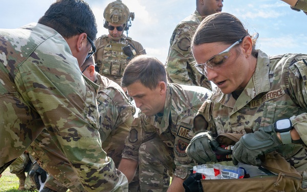 Multiple U.S. Air Force personnel in OCP (Operational Camouflage Pattern) uniforms are performing tactical combat casualty care (TCCC) on a simulated casualty lying on grass. At least two individuals are visibly identified as medics, with one female medic on the right accessing a medical kit. This appears to be a training exercise based on the controlled environment and observable simulation indicators.