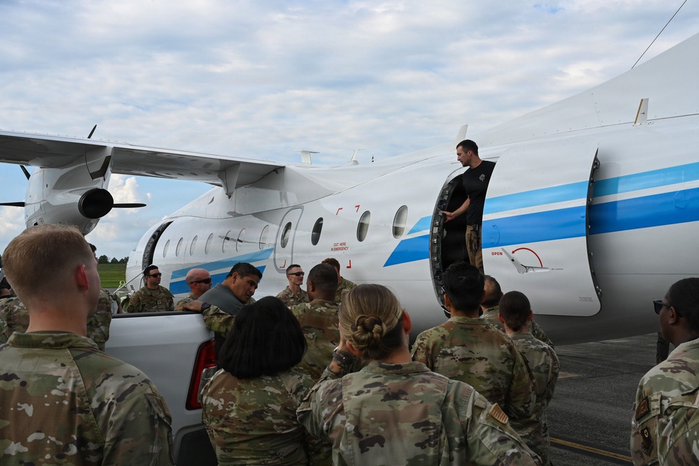 A group of military personnel in camouflage uniforms are gathered on a tarmac around a white and blue twin-turboprop aircraft. A civilian instructor or demonstrator in black clothing stands in the open doorway of the aircraft, apparently conducting a briefing or training exercise. The scene suggests an aircraft familiarization or emergency procedures training event.