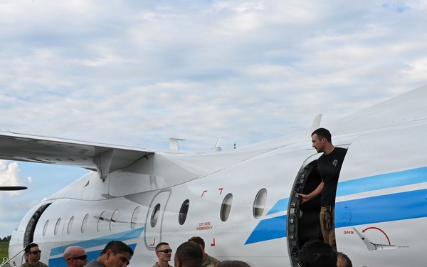 A group of military personnel in camouflage uniforms are gathered on a tarmac around a white and blue twin-turboprop aircraft. A civilian instructor or demonstrator in black clothing stands in the open doorway of the aircraft, apparently conducting a briefing or training exercise. The scene suggests an aircraft familiarization or emergency procedures training event.
