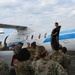 A group of military personnel in camouflage uniforms are gathered on a tarmac around a white and blue twin-turboprop aircraft. A civilian instructor or demonstrator in black clothing stands in the open doorway of the aircraft, apparently conducting a briefing or training exercise. The scene suggests an aircraft familiarization or emergency procedures training event.